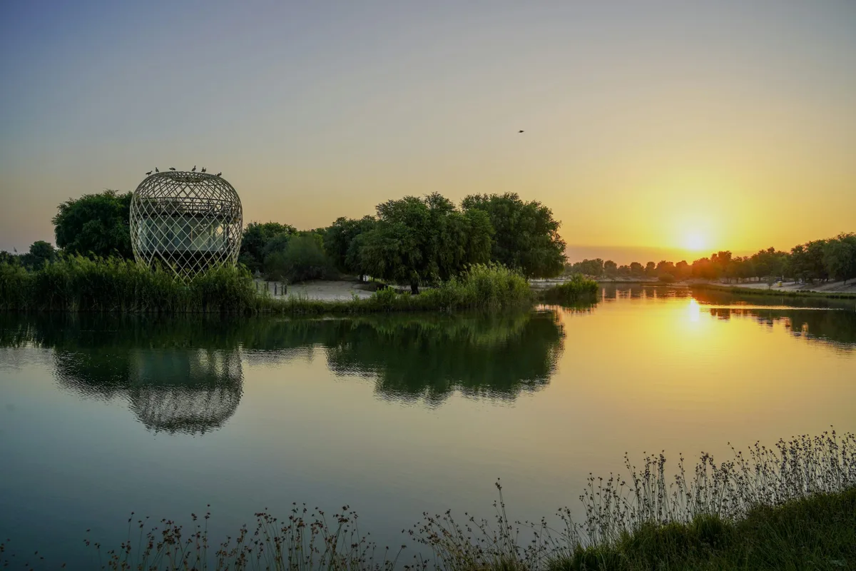 Sunset over Love Lake in Dubai, with trees and a sculpture beside the water